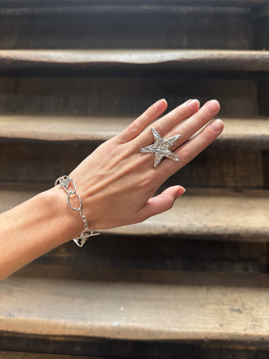 A hand wearing a silver starfish ring and a silver link bracelet, posed against rustic wooden stairs.
