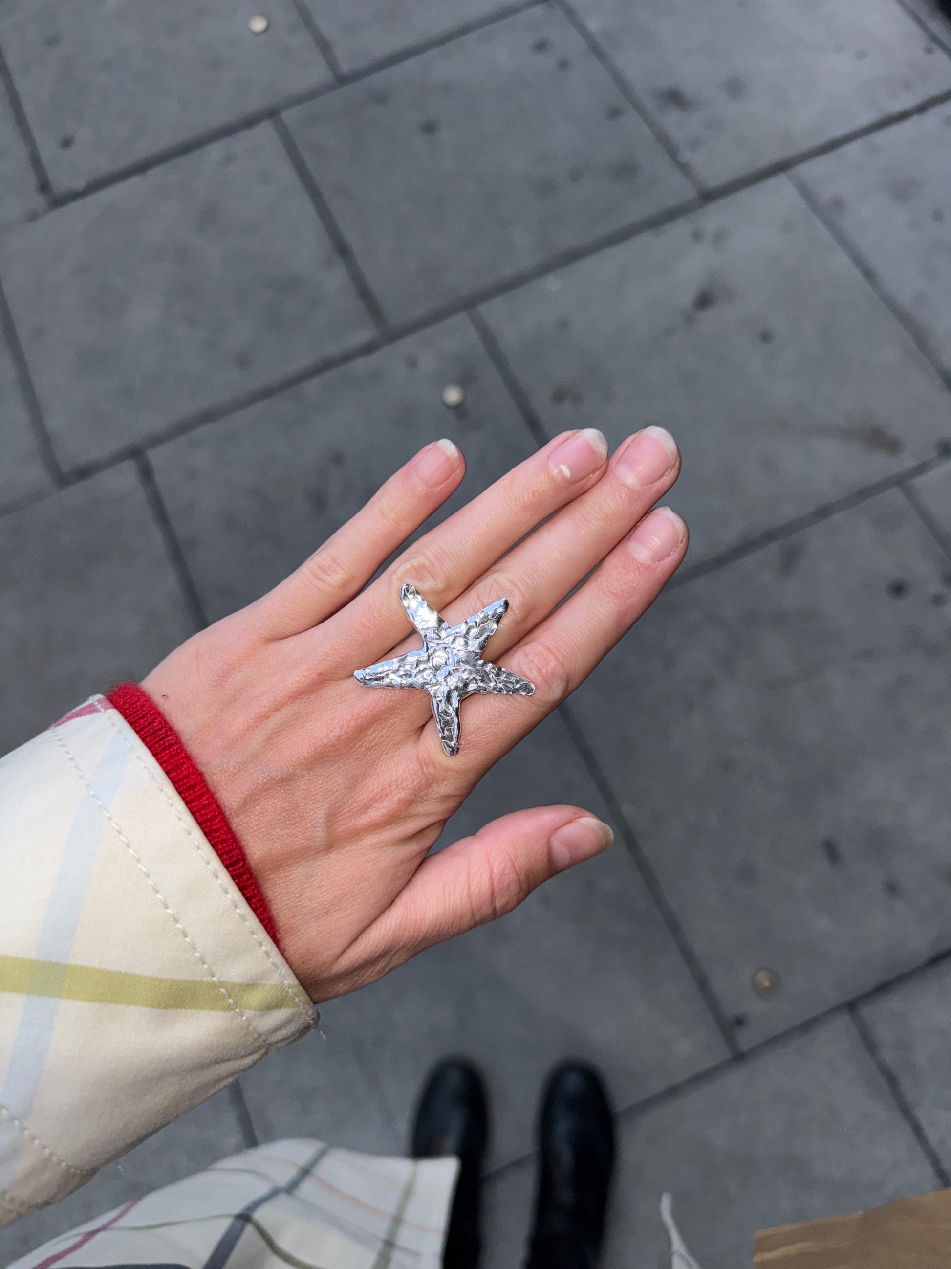 A hand wearing a silver-toned starfish ring, photographed outdoors on a gray paved surface with part of a person's coat and shoes visible.