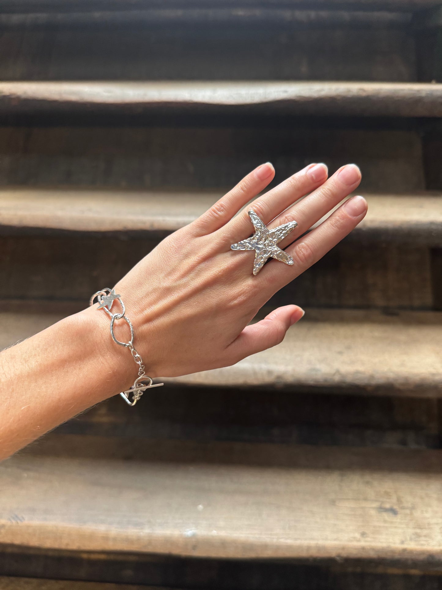 A hand wearing a silver starfish ring and a silver link bracelet, posed against rustic wooden stairs.