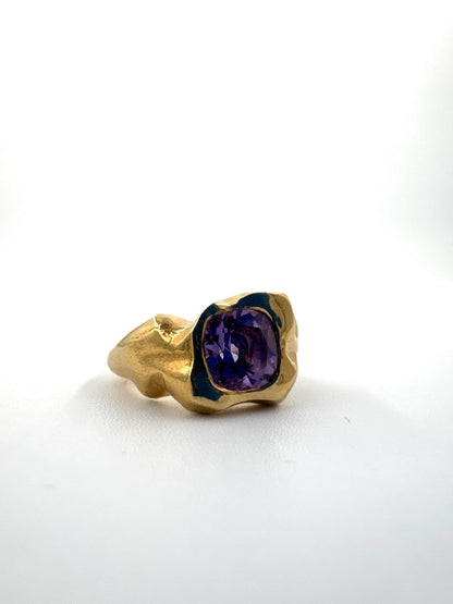 A gold ring with a textured, organic-shaped setting holding a cushion-cut purple gemstone, photographed on a clean white background.